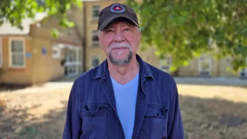 Stuart Woodward/BBC A man wearing a sun hat and a navy jacket with a blue shirt and he is looking at the camera with a faint smile.