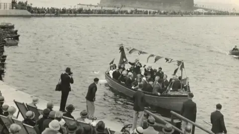 Wirral Archives A boat carrying a number of passengers is sailing across the water at the New Brighton Marine Lake in this black and white photograph. A number of spectators watch - some in deckchairs while others are standing.