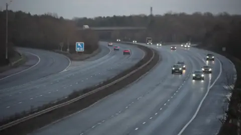 The M62 motorway with the east and westbound carriageways separated by a strip of grass and metal central reservation barrier. A number of vehicles can be seen on both sides of the road and have their headlights turned on. Trees and a bridge are visible in the distance along with a blue road sign.