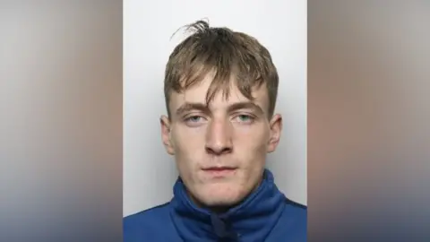 Police custody image of Blake Lester, a man with brown hair, wearing a blue top and staring straight at the camera.