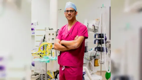 Carla Mabb A doctor wearing pink scrubs standing in an operating theatre with his arms folded