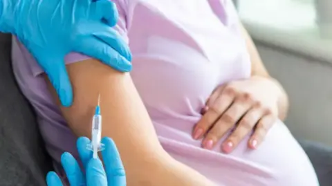 A pregnant lady being administered a vaccine by a medical professional wearing blue gloves. She is wearing a pink t-shirt and has her left hand draped over her stomach.