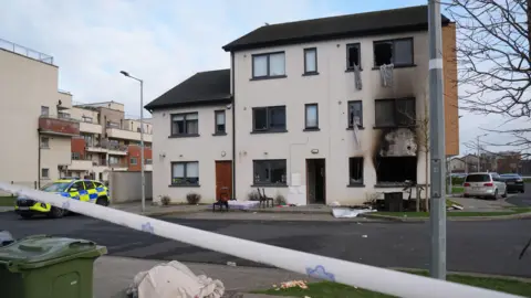 PA Media Two burnt out windows sit at the end of a row of three white terrace houses. There is black rubble and burnt household objects sitting outside on the green grass at the foot of the window. A single leafless tree sits in front of the objects on the grass. The other two houses are intact. A blue and yellow police car sits to the left of the houses. Blue and white police tape sits in front of the tarmac road which separates the tape from the houses. 