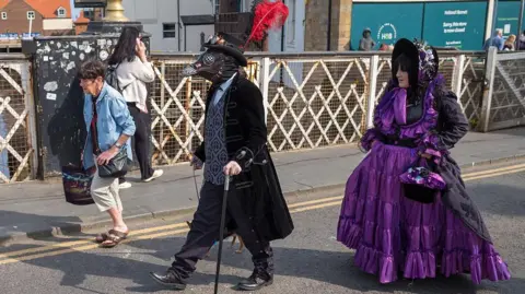 Getty Images Three people walking from left to right across a bridge dominate the foreground. The central person is wearing a theatrical costume which includes a long black coat, dark trousers, tall hat, black boots and a bird-like mask with a long beak shape and circular eye pieces. The woman on the right wears a voluminous purple dress with multiple ruffled layers, a structured bodice and a wide skirt. The person on the left wears everyday clothing: a light brown jacket and light-colored cropped trousers.
