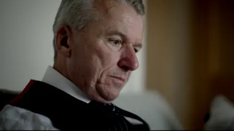Firecrest Films A man wearing a white shirt, black waistcoat and black tie looks down at some paperwork in an office setting