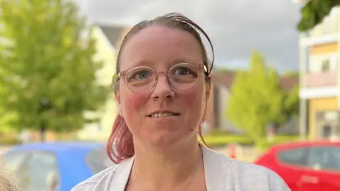 Becki Longstaff has shoulder length hair tied back. She is stood in a car parking in front of blue and red cars, also is also wearing glasses.