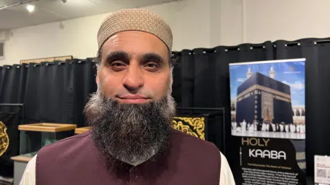 Amanda White/BBC A man wearing a light brown hat and a maroon vest stands in front of a black curtain and a large poster featuring the Kaaba in Mecca. Display cases are also visible in the background.