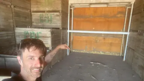 Luke Abblitt Luke, a man, with short brown hair. He is wearing a black T-shirt and is pointing towards a large empty space inside a storage shed.