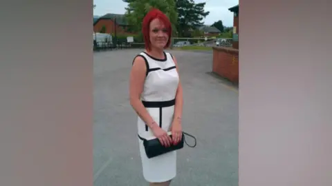 Tiffany Render has dyed-red hair and is smiling. She is standing near a building, dressed in a smart long white and black dress holding a black handbag.