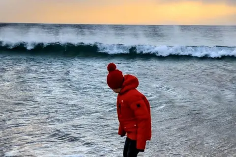 Gareth Taylor A boy wearing a red jacket and red bobble hat standing in the sea. Waves are crashing into the background