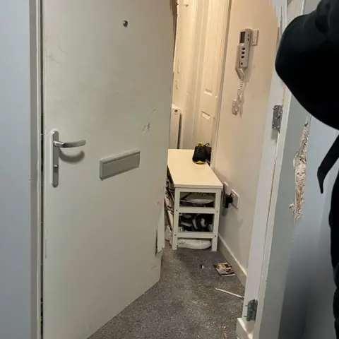 A smashed white door to a flat with debris on the floor