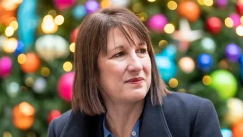 Getty Images Chancellor Rachel Reeves in navy blue coat and blue shirt photographed in front of colourful Christmas tree decorations at Leeds Corn Exchange