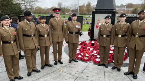 A line of eight servicemen wearing brown uniforms and black belts, standing to attention beside a cenotaph and posing for a picture. There are lots of poppy wreaths laying at its base.