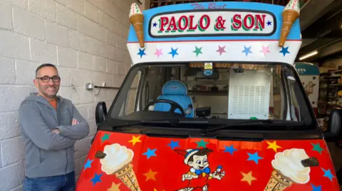 A man stands alongside a brightly coloured ice cream van, with pictures of ice creams and a cartoon character on the front, and the nams Paolo and Son across the top