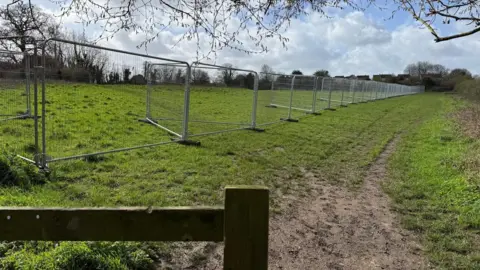 People for Packsaddle A temporary metal fence stretches into the distance, dividing a muddy path from a green field.