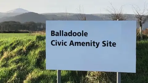 A white sign in front of mountains that reads Balladoole Civic Amenity Site