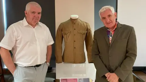 BBC/Sofia Luis-Hobbs Two older men stand next to a display case with an old-fashioned military jacket in it.