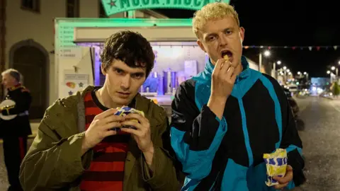 Two men are standing in front of a chip van at night time. The man on the left is about to take a bite of a burger in a yellow and blue paper bag. The man on the right is eating a chip from a blue and yellow paper bag. The man on the left has short brown hair and is wearing a brown coat and red t shirt. The man on the right has short blonde hair and is wearing a blue and black tracksuit top.