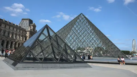 Mohammed Badra/EPA/Shutterstock Two modern glass pyramids at the Louvre in Paris with an older building to the left. The sky is blue and there are a few people sitting in front of the pyramids. There is a ferris wheel in the distance on the right. 