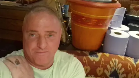 Supplied Raymond McDonald is a man in his fifties with a receding hairline. He is sitting down on a brown and cream patterned sofa. He is wearing a light green t-shirt and has his hand on his shoulder. There is a tattoo on one finger where a ring would be. Behind him is a flower pot and a number of rolls of blue cleaning paper.