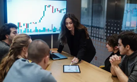 Getty Images Woman points at financial graph on tablet as colleagues in meeting look on