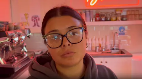 Adrian Horms/BBC, a woman wearing a hoodie stood in a red-light room. A neon sits in the sign background reading "Flower",