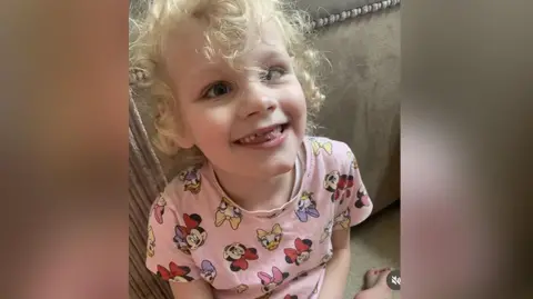 Seven-year-old Isla with blond curly hair wearing a pink Disney patterned t-shirt sits on the floor leaning on a beige couch. She is smiling.