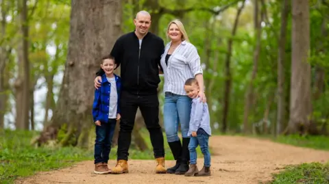 Brain Tumour Research Charlie and Siobhan pose for a photo on a forest path with their two young sons. Charlie has a shaved head and wears a black zip up top, black jeans and brown boots. Siobhan has long blonde hair and wears a grey striped shirt, jeans, and black boots. The boys wear t shirts with open shirts over the top, jeans, and boots.