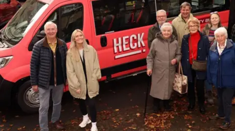 A group of eight elderly people stand in front of a pink mini-bus with the word HECK written on its side