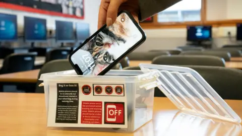 Image shows a mobile phone being put inside a clear plastic box in a classroom.