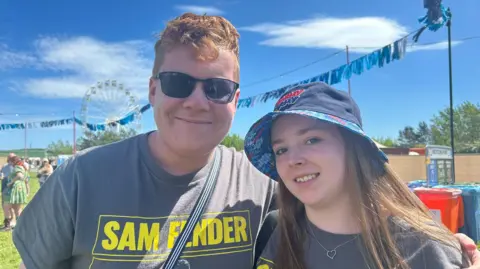 A man and woman wearing an yellow and grey Sam Fender t-shirt smile at the camera on a music festival site.