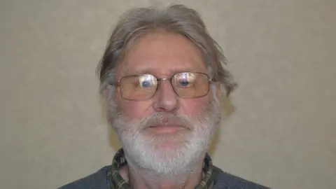 Contributed A man with grey hair that has grown down to his ears. He also has a thick grey beard. He is wearing glasses along with a grey jumper and chequered green shirt.