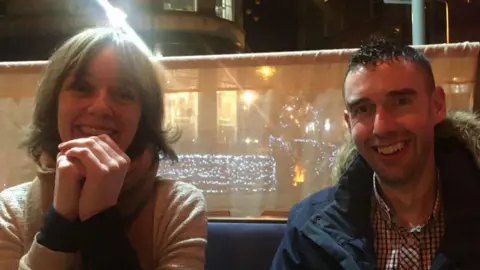 Dawn Sincock (left) sitting next to her brother, David. It is night-time and the area they are seated at is lit up. Both are smiling into the camera. Dawn has dark hair and is wearing a beige top and scarf. David has short dark hair, a checked shirt and a navy blue parka jacket with a fur-style trim.