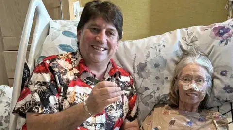 John Owen John and Rowena Owen sit in a large hospital type bed. Rowena has a tube going into her nose, held in place by micropore tape. She is wearing a top with cats on while John wears a shirt with cats on. The are both smiling. Rowena is looking into camera but John is looking slightly off camera.