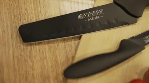 Two knives with black blades and handles on a chopping board with rounded tips and the logo 'Viners Assure'