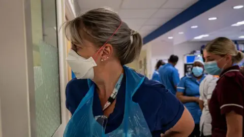 Getty Images A medic standing in what looks like the emergency room of an NHS hospital in Covid. She is staring through a window perhaps looking outside. She is wearing a plastic blue apron over her scrubs and has a white masks on. Behind her are other members of staff wearing PPE. 