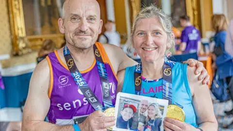 Claire Reece/Sense Claire Reece and Steve Childerley after running the London Marathon. They are wearing bright coloured vests, with medals around their necks and are smiling at the camera. They holding a picture of themselves with Reece in front. They are in a grand room with lage gilded mirrors and people milling around. 