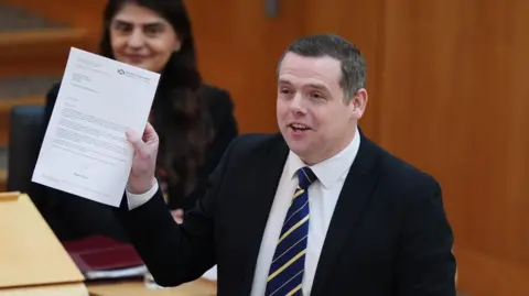 PA Media Douglas Ross addressing MSPs in the Scottish Parliament chamber. He is holding up a letter on Scottish government paper and speaking. 