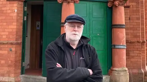 Leslie Holmes, culture and heritage projects manager at the Salford Lads and Girls Club stands outside the club. He is wearing a black cap and black top and fleece and has grey hair and beard. 
