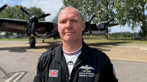 Un hombre sonríe a la cámara. Lleva una camiseta blanca debajo de un mono negro con la bandera de la Unión y el logotipo de la RAF. Está de pie en una pista asfaltada con un bombardero Lancaster al fondo. También se ven árboles y un cielo azul.