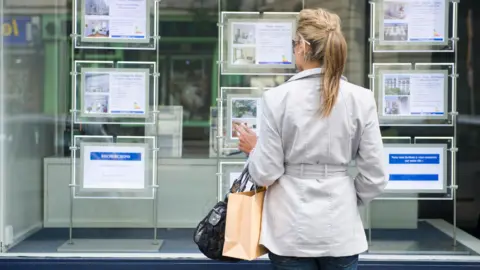 A woman with a blonde ponytail and a light grey trench coat holds a handbag and a paper bag while looking in a real estate window at real estate listings.
