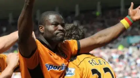 Getty Images Footballer in an orange short-sleeved Wolves shirt with a wristband showing the yellow, red and green colours of Cameroon accepts the acclaim of supporters after scoring a goal