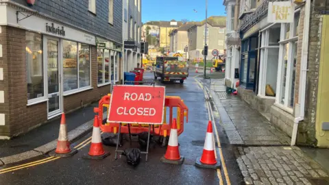 A small street. There is a red road closed sign. There are cones. In the background is a truck and vans parked. There are buildings around the road. The sky is blue. 