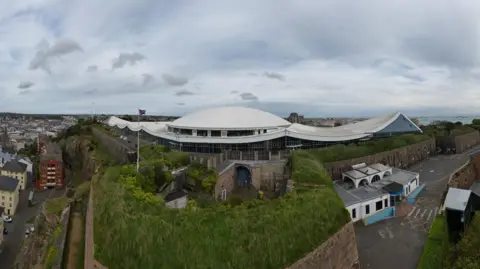 BBC A wide shot of the Fort Regent leisure centre. It is a large white-roofed building surrounded by trees. There is a green hedges and grass around it along with solid stone walls and lots of houses. 