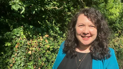 Amy Holmes/BBC A picture of Lib Dem MP Victoria Collins, who is MP for Harpenden and Berkhamsted. She has curly dark shoulder length hair and is wearing a magenta green suit jacket. She is stood in front of a green bush.