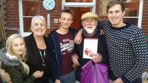 Collect/PA Real Life Gabe's sister Miffy, grandmother Marie, Gabe, grandfather Peter and older brother Harry - all standing smiling at the camera. They are standing outside of a house. Grandfather peter is holding a purple carrier bag and wearing a flatcap.