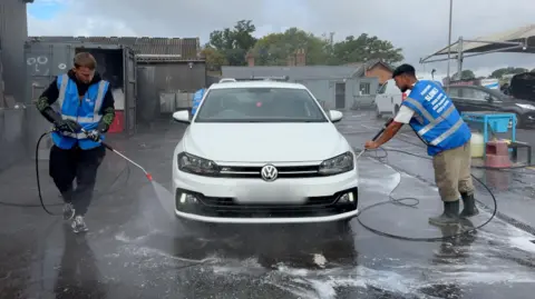 Image shows a white Volkswagen Polo in the centre. It is being washed. Either side of the car is a worker holding a jet wash. They are washing off the tyres. The worker on the left is wearing a black tracksuit and a blue high-vis. The worker on the right is wearing beige trousers and a white top. He is also wearing a blue high-vis. Behind the car are two grey buildings. 