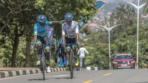 Two cyclists, one wearing a blue jersey and black shorts and the other in a white jersey and black shorts ride along a tree-lined street in Kigali while a maroon car is seen in the background