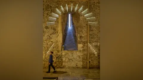 David Levene/Art Fund/PA Media A slotted window in the interior wall of Norwich Castle, framed by lots of glass lights hanging down. Someone strides past.