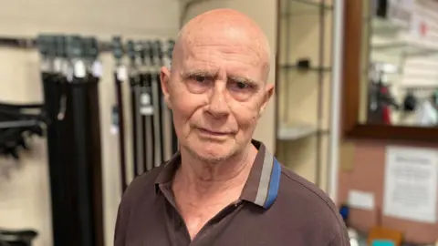 Sam Read/BBC Colin Gilmour in a brown polo shirt stands against the backdrop of belts inside the menswear store.
